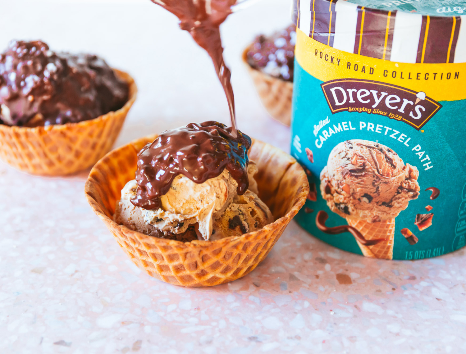 Photo of a scoop of Dreyer's Carmel Pretzel Path ice cream in a waffle cone cup, drizzled with chocolate.